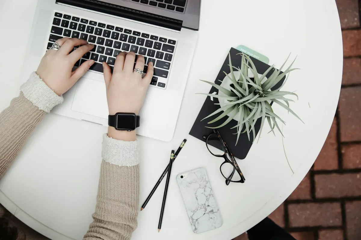Freelancer de tráfego pago digitando em notebook sobre mesa branca com planta, óculos e celular ao lado.
