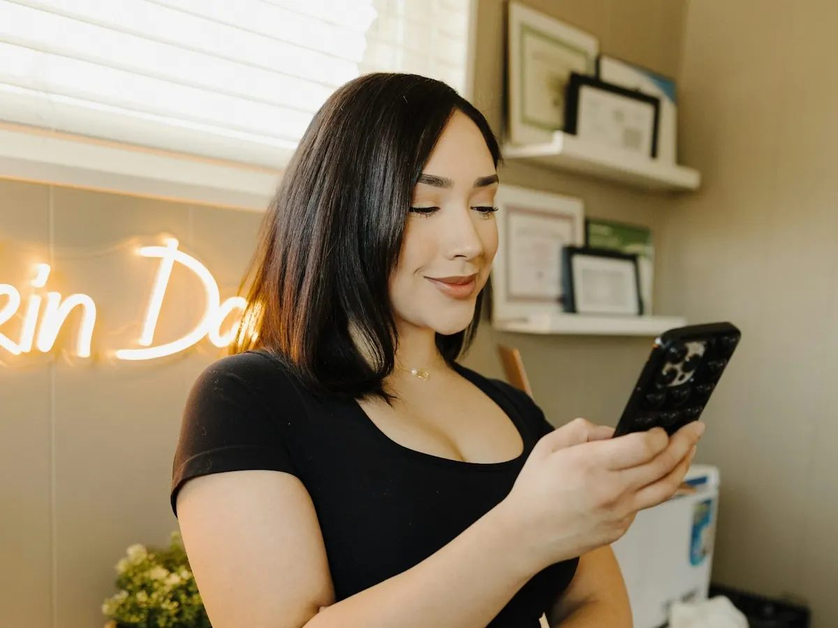 Dona de uma pequena loja sorrindo enquanto responde mensagens no celular, com prateleiras e um letreiro de neon ao fundo.