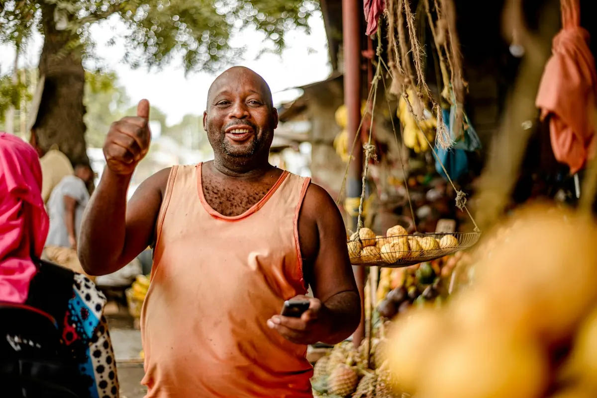 Feirante sorrindo e fazendo sinal de positivo com o celular na mão, ao lado da sua banca de frutas.