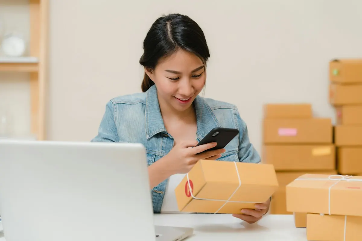 Empreendedora sorrindo com o celular na mão ao lado do notebook e de caixas de papelão prontas para envio.