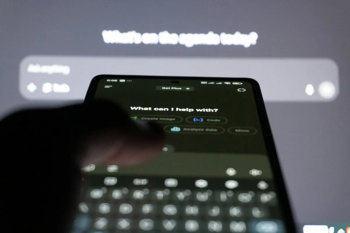 Mão digitando em smartphone com interface de assistente de IA, e monitor ao fundo com tela de chatbot.
