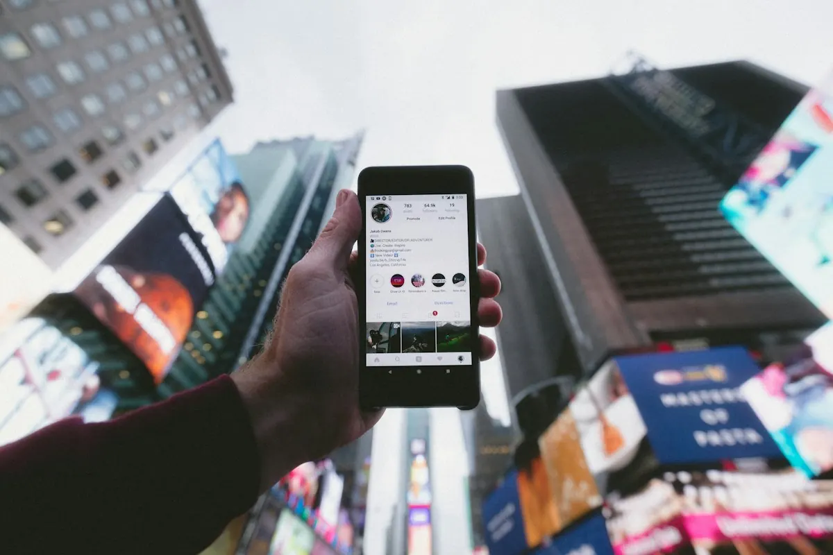 Mão segurando um celular com um perfil do Instagram aberto em meio a prédios e painéis da cidade.
