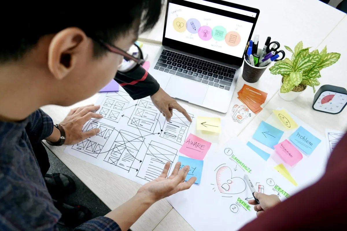 Pessoa desenhando wireframes de páginas em papel com post-its coloridos, ao lado de um notebook.