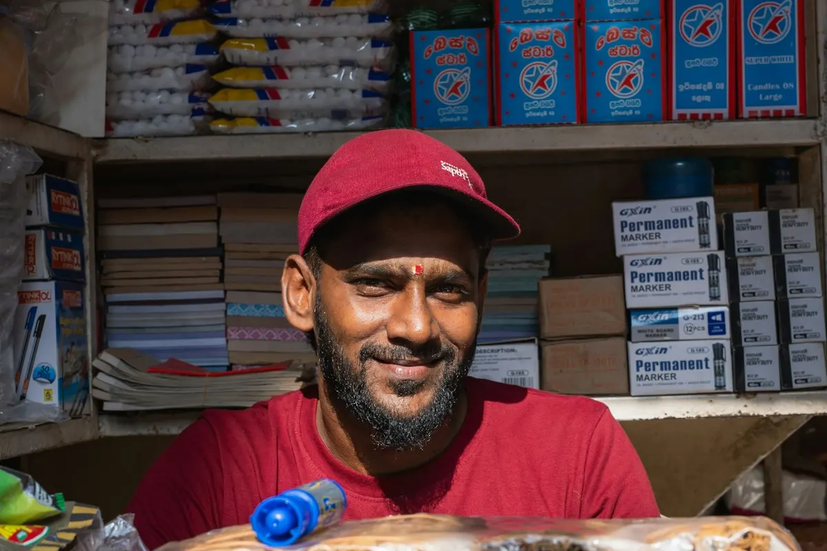 Comerciante de boné vermelho sorrindo atrás do balcão de uma pequena loja de bairro, com prateleiras cheias de produtos ao fundo.