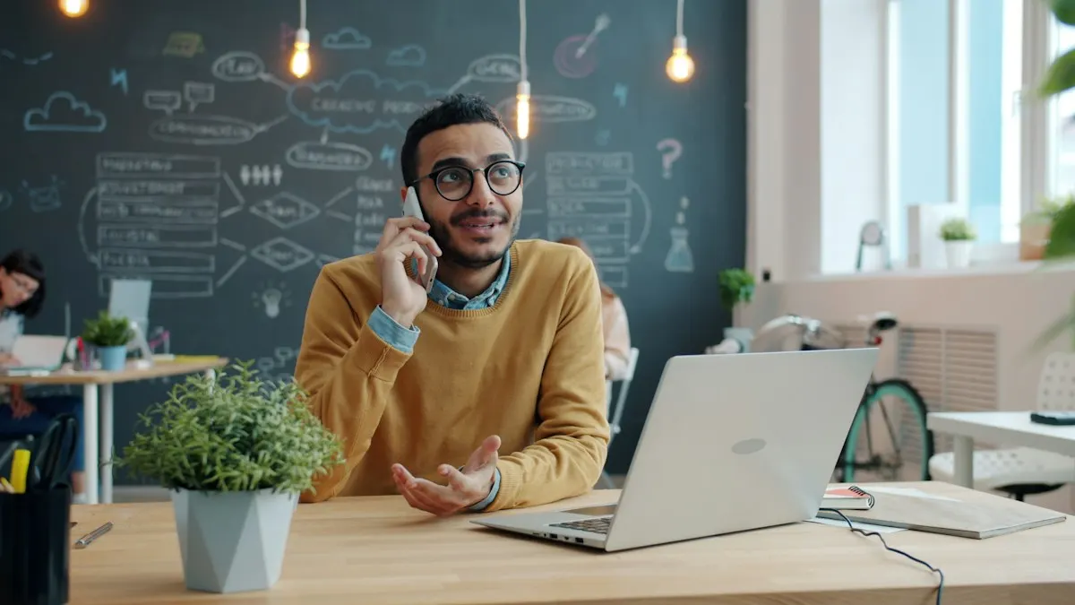 Homem de óculos e suéter amarelo falando ao celular na mesa de trabalho, com notebook aberto, atendendo uma ligação de cliente.