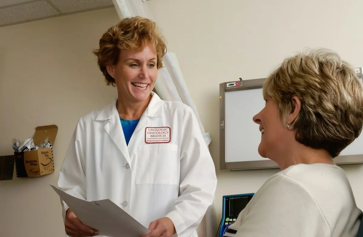 Médica sorridente de jaleco branco conversando com uma paciente dentro de um consultório.
