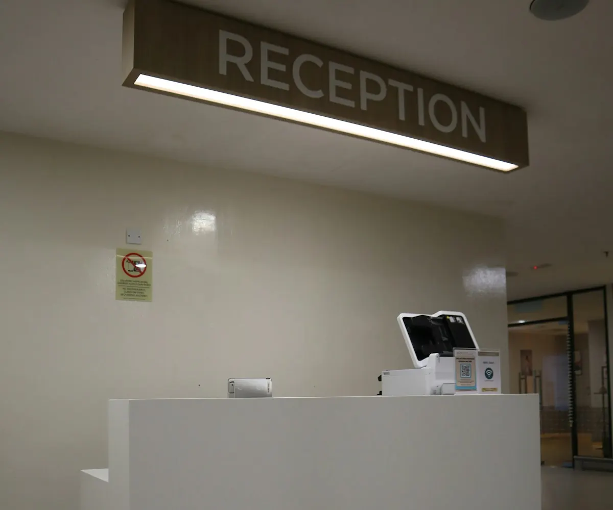 Balcão branco de recepção de uma clínica com a placa Reception iluminada no alto.