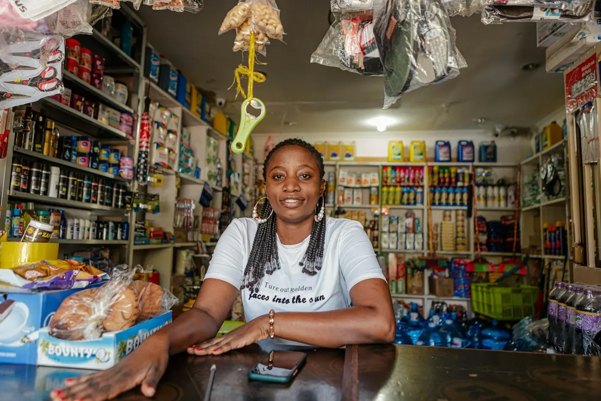 Dona de uma pequena loja sorrindo atrás do balcão, com prateleiras cheias de produtos ao fundo e o celular sobre o balcão.