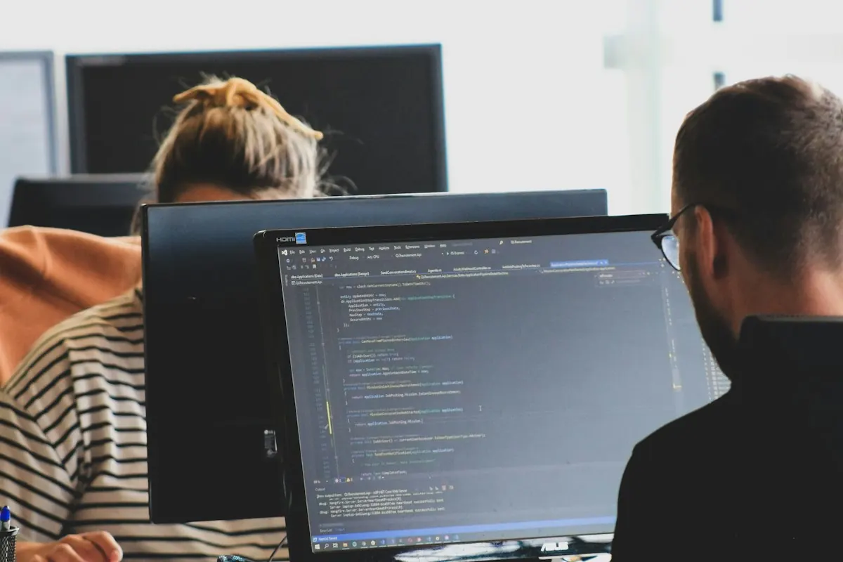 Duas pessoas programando em monitores num escritório claro, com linhas de código na tela.