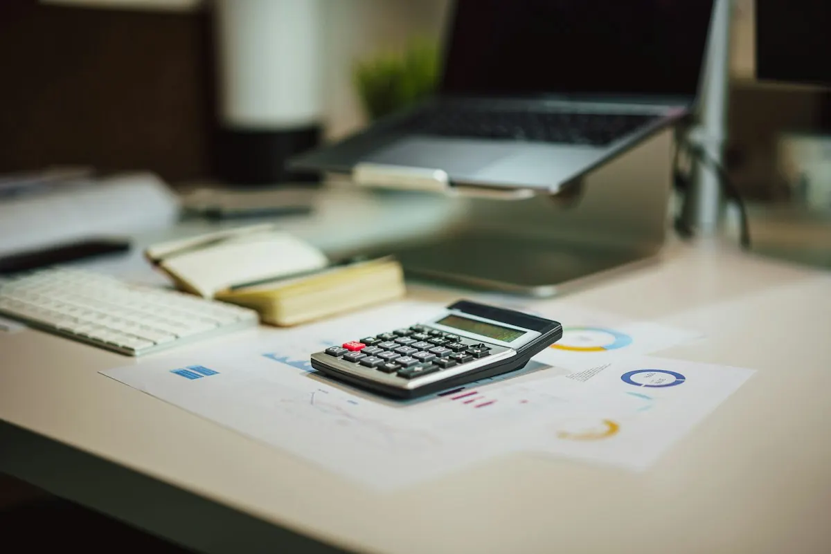 Calculadora sobre relatórios impressos com gráficos, ao lado de um notebook e de um teclado em uma mesa de escritório.