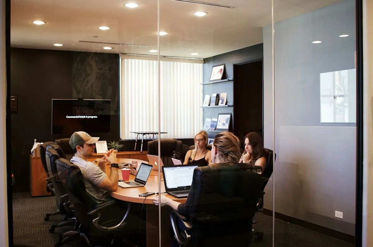 Equipe reunida em volta de uma mesa com notebooks em uma sala de reuniões envidraçada.