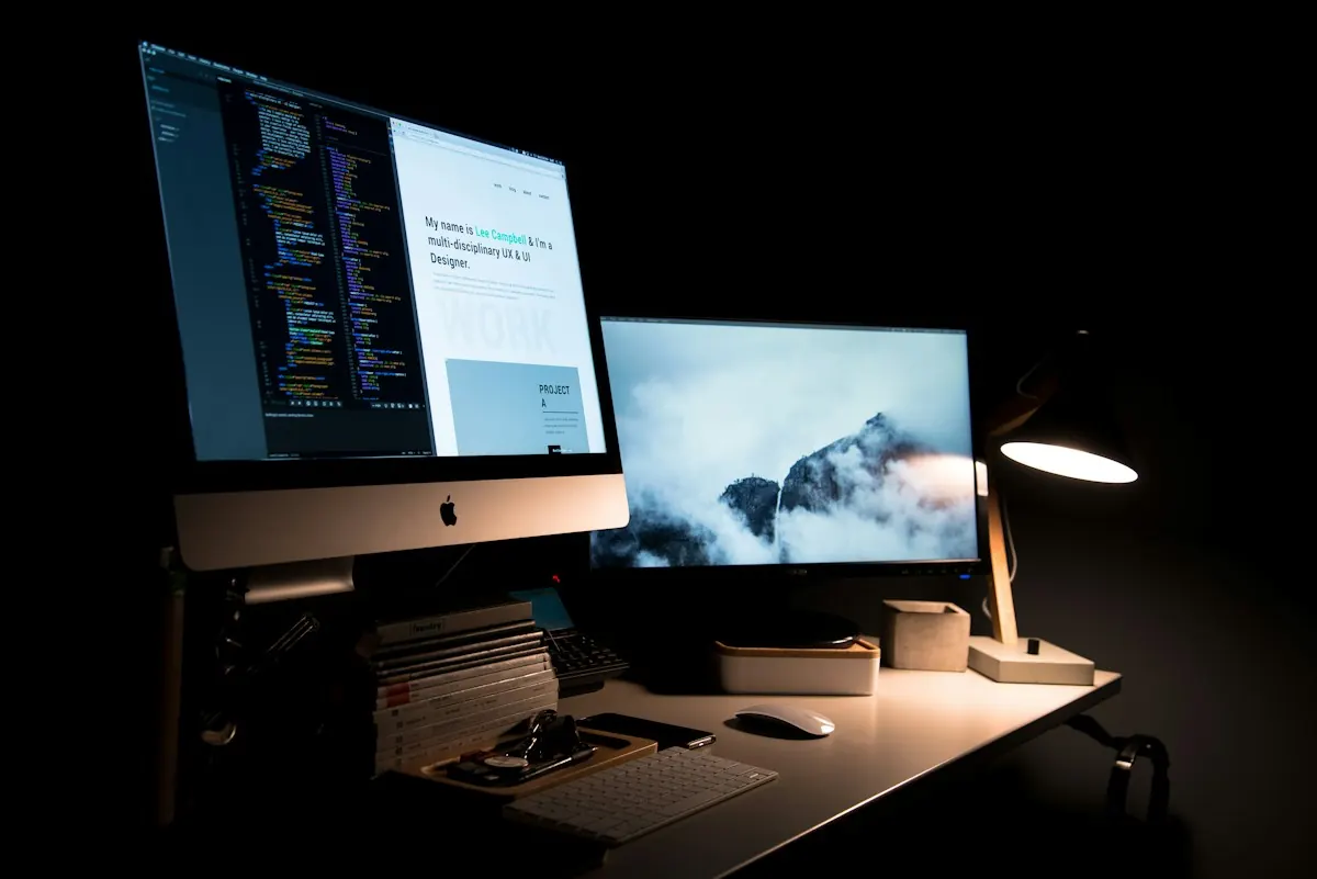 Mesa de trabalho com iMac exibindo código e preview de site, segundo monitor e luminária em ambiente escuro.