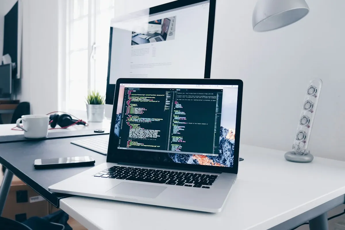 Laptop com editor de código em primeiro plano e monitor ao fundo exibindo um site em desenvolvimento.