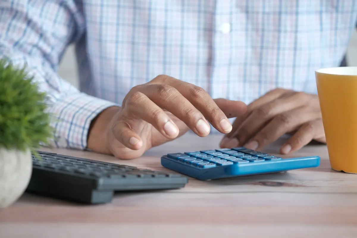Pessoa de camisa xadrez fazendo contas numa calculadora sobre a mesa, ao lado de um teclado e de uma caneca, planejando o orçamento de marketing.