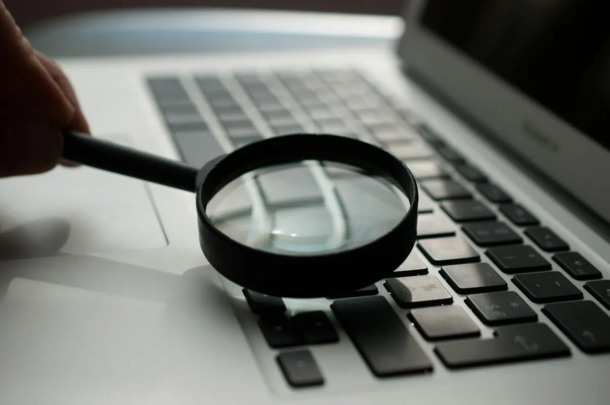 Mão segurando uma lupa sobre o teclado de um notebook prateado, como quem examina de perto os resultados de uma busca.