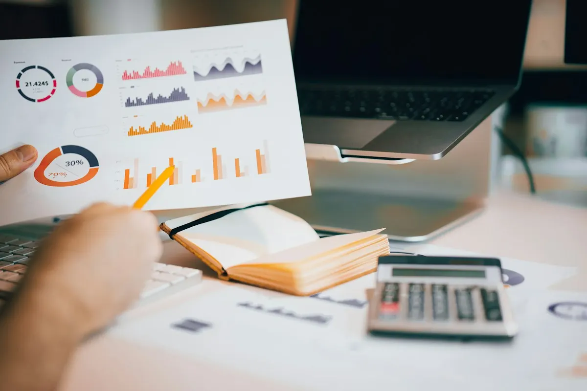 Pessoa segurando relatório impresso com gráficos de resultados de marketing sobre a mesa, ao lado de calculadora e notebook.