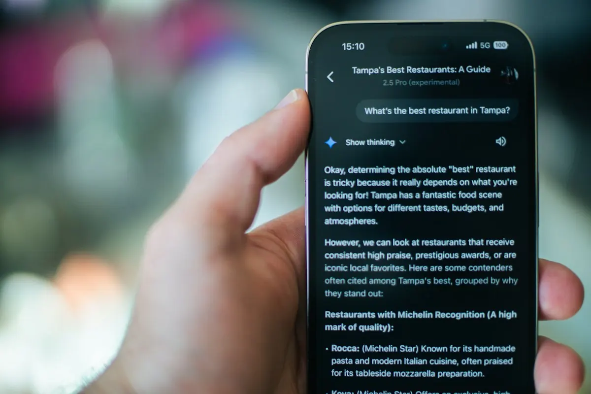 Mão segurando um celular em que um assistente de IA responde à pergunta sobre qual é o melhor restaurante da cidade.