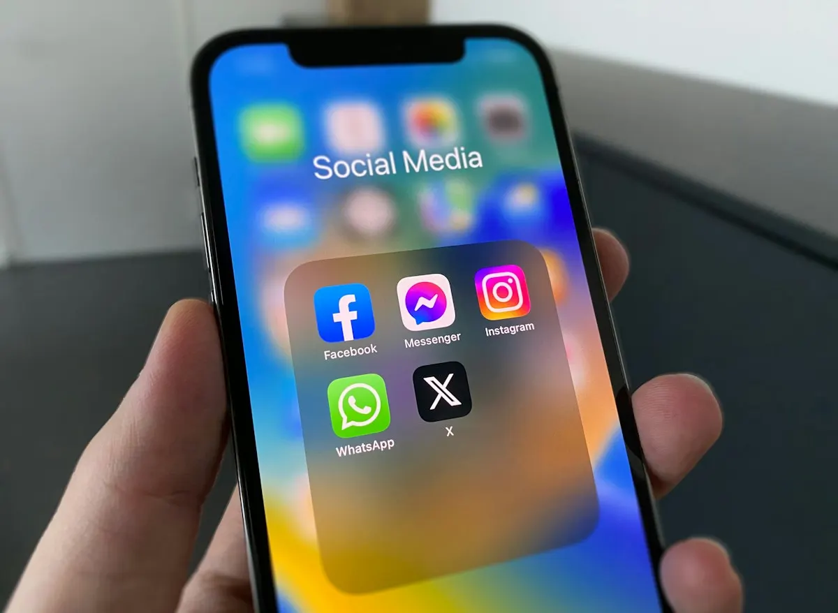 Mão segurando um iPhone com uma pasta de aplicativos de redes sociais aberta, mostrando os ícones do Facebook, Messenger, Instagram, WhatsApp e X.