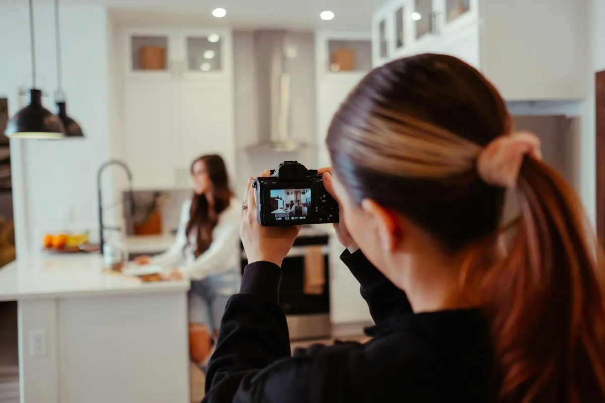 Mulher de costas segurando uma câmera fotográfica e gravando outra mulher que trabalha numa cozinha clara, produzindo conteúdo para redes sociais.