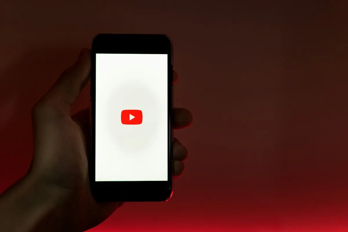 Mão segurando um celular com o logotipo do YouTube na tela, sobre um fundo vermelho.